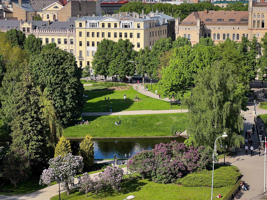 Aerial view of a green park with people walking and sitting on benches, surrounded by trees and blooming flowers, next to a small pond. Historic buildings are visible in the background.