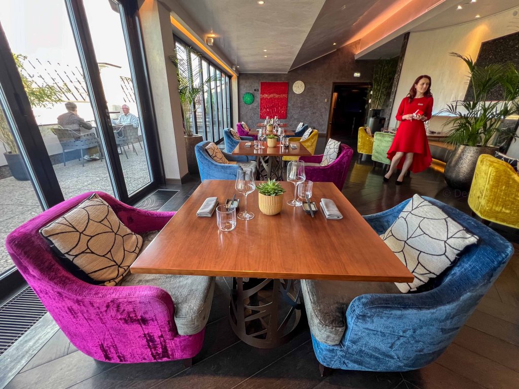 A modern restaurant interior featuring colorful plush chairs in pink, blue, and yellow, with a wooden dining table set for two. A woman in a red dress is seen walking in the foreground, while other diners enjoy their meals in the background by large windows with a view.