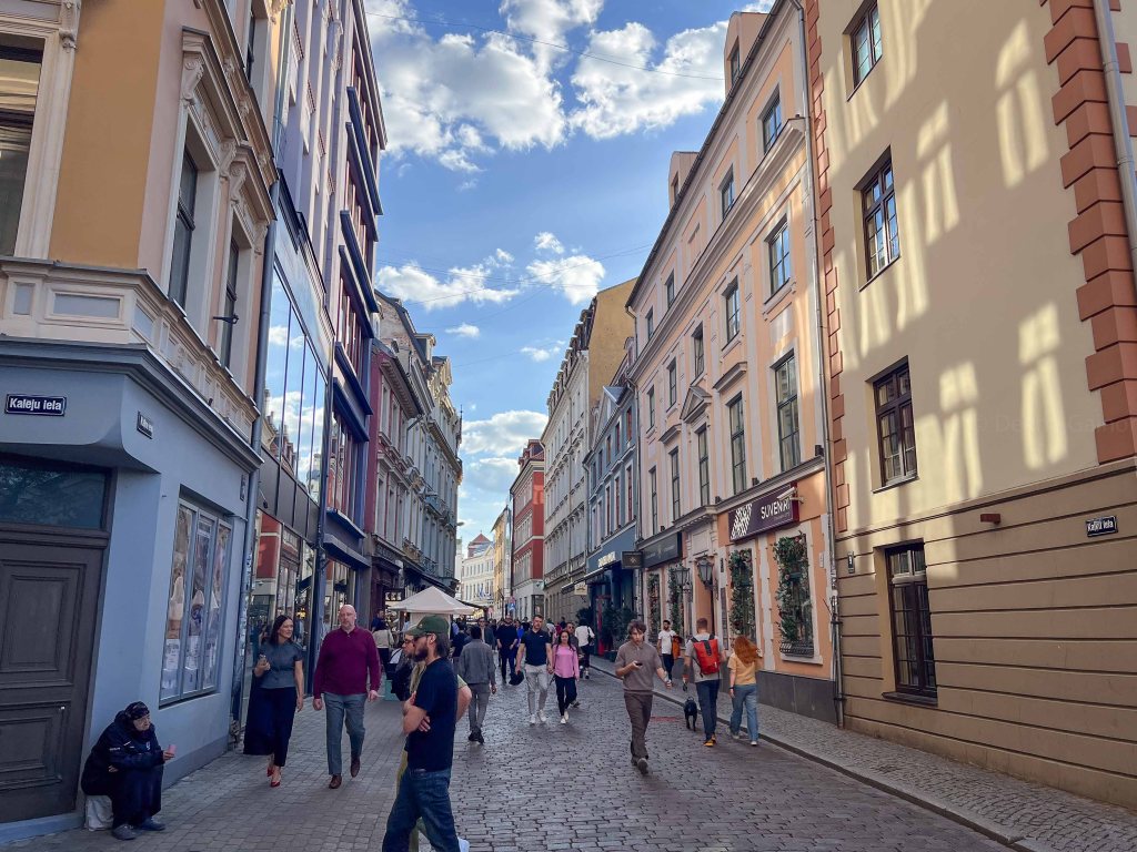 A bustling street scene in a city with colorful historical buildings and people walking, some shopping and socializing under a partly cloudy sky.