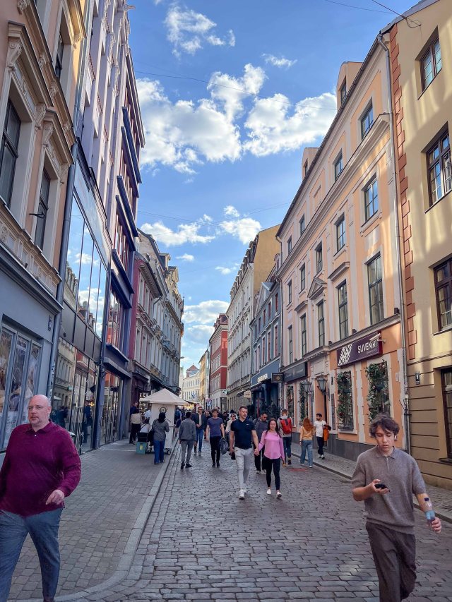 A busy cobblestone street lined with colorful buildings, featuring pedestrians walking, a clear blue sky with scattered clouds, and storefronts on either side.
