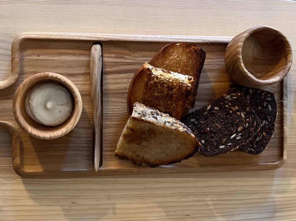 A wooden serving board featuring slices of different types of bread, including toasted white bread and dark, seeded bread, accompanied by a small bowl of dip.