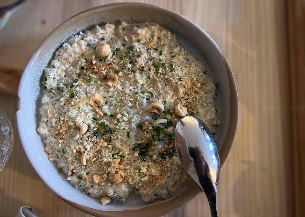 A bowl of creamy dish topped with chopped hazelnuts, herbs, and breadcrumbs, with a spoon resting beside it.