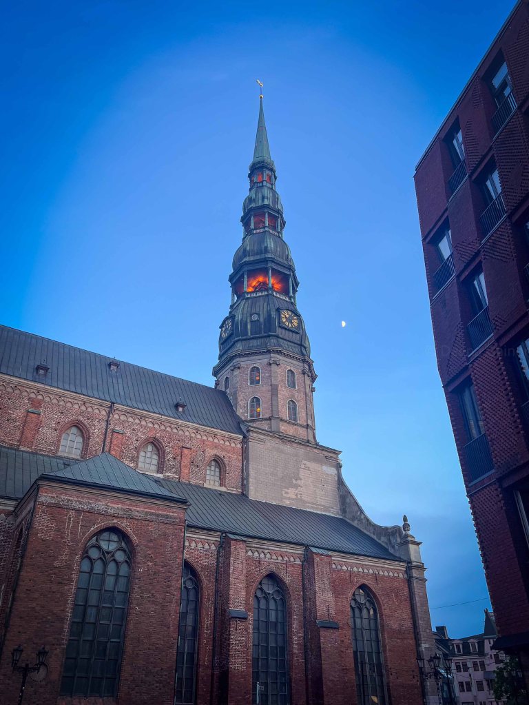 A view of a tall church spire against a twilight sky, with intricate brickwork and large arched windows visible.