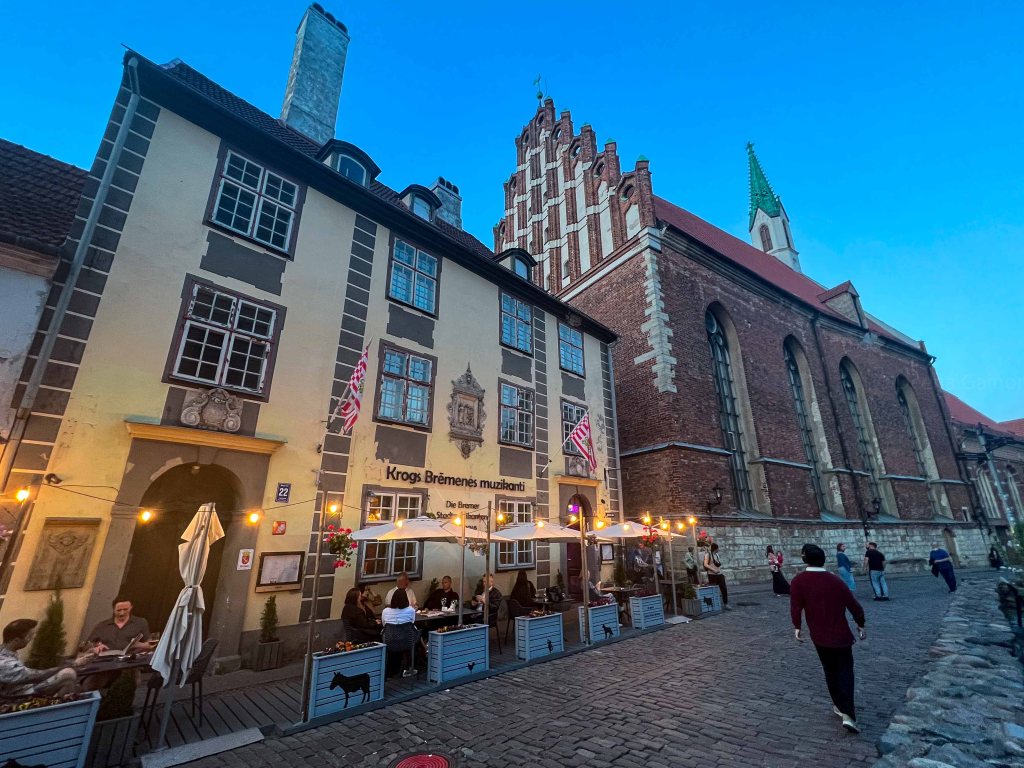 A street view of a quaint restaurant with outdoor seating and string lights, located next to an old brick church with a steeple, under a twilight sky.