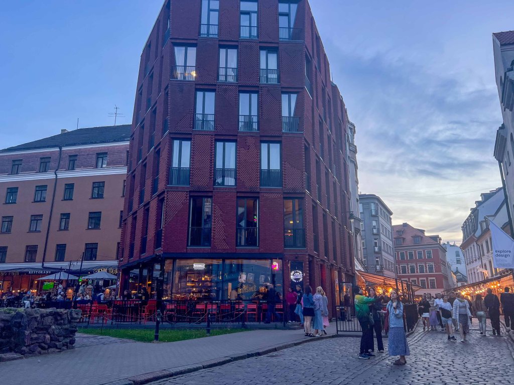 Street view of a modern red brick building with large windows, surrounded by traditional architecture and people dining at outdoor cafes during sunset.