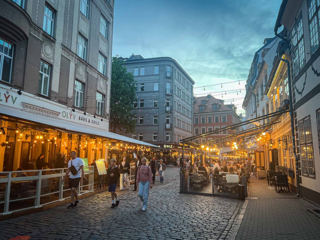 A bustling cobblestone street lined with restaurants and cafes, illuminated by string lights during twilight.