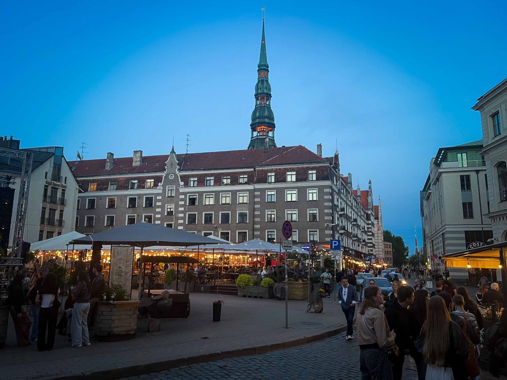 Evening view of a bustling square with outdoor dining, featuring historical buildings and a tall, pointed tower against a blue sky.