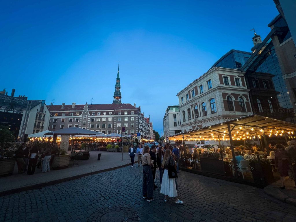 A vibrant evening scene at a bustling market square with people socializing, outdoor dining setups, and historic buildings illuminated under a blue sky.