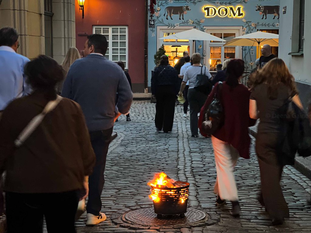 A lively street scene with people walking along a cobblestone path, passing by a restaurant named 'DOM' with outdoor seating and decorative lights. A small fire burns in a metal basket on the sidewalk.