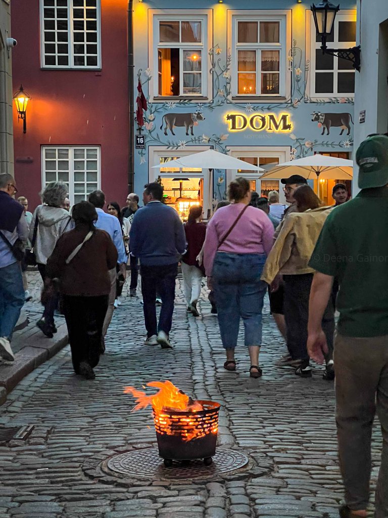 A cobblestone street bustling with pedestrians in the evening, featuring a brazier with flames burning in the foreground and a restaurant named 'DOM' in the background, illuminated with warm lights.