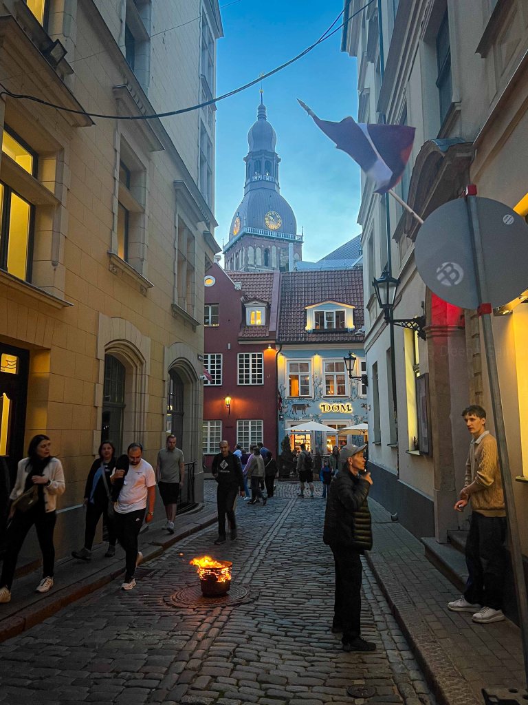 A cobblestone street in the evening with people walking. A fire in a metal container is visible in the foreground, and a prominent church steeple is seen in the background. Several shops and buildings line the street.