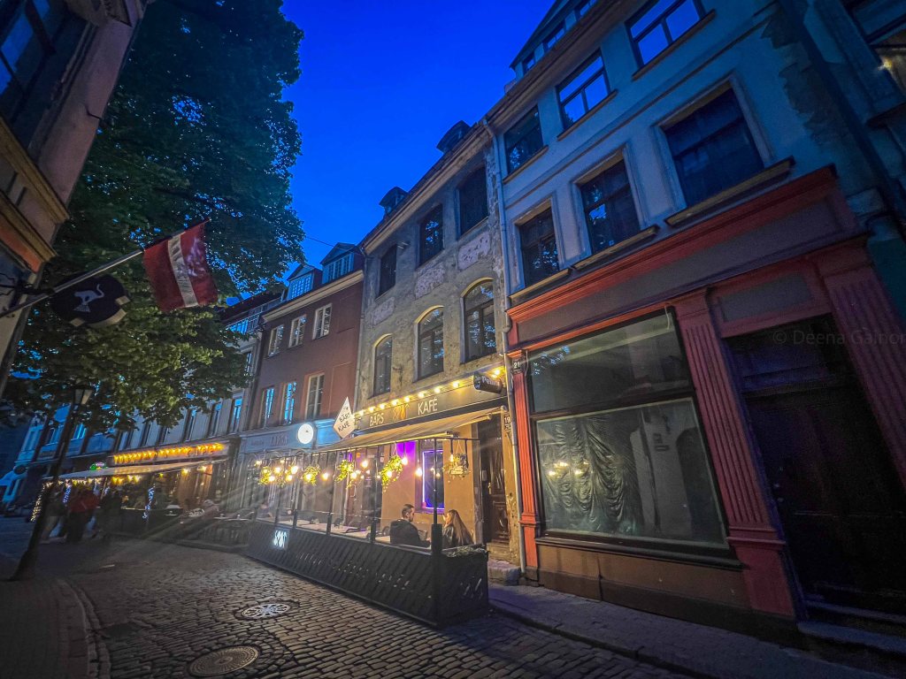 A cobblestone street in a historic area, illuminated by warm lights from outdoor cafes and restaurants, with trees and Latvian flags in the foreground against a twilight sky.