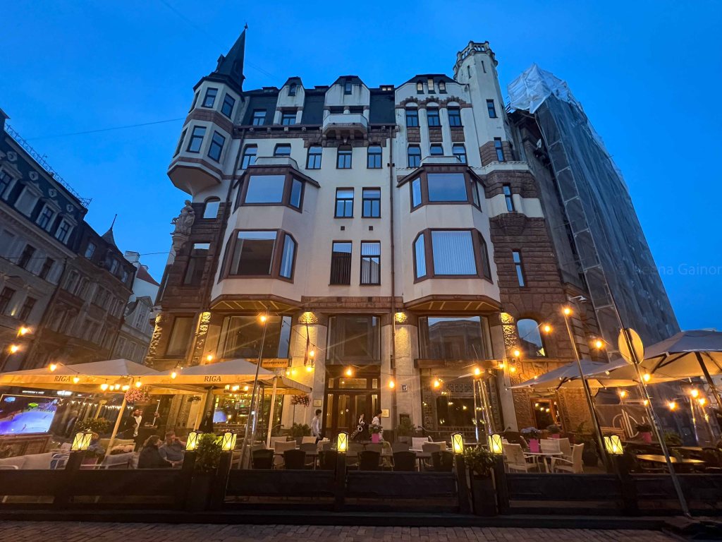Exterior view of a stylish building with multiple windows and a turret, illuminated at dusk, featuring a lively outdoor café with bright lights and umbrellas.