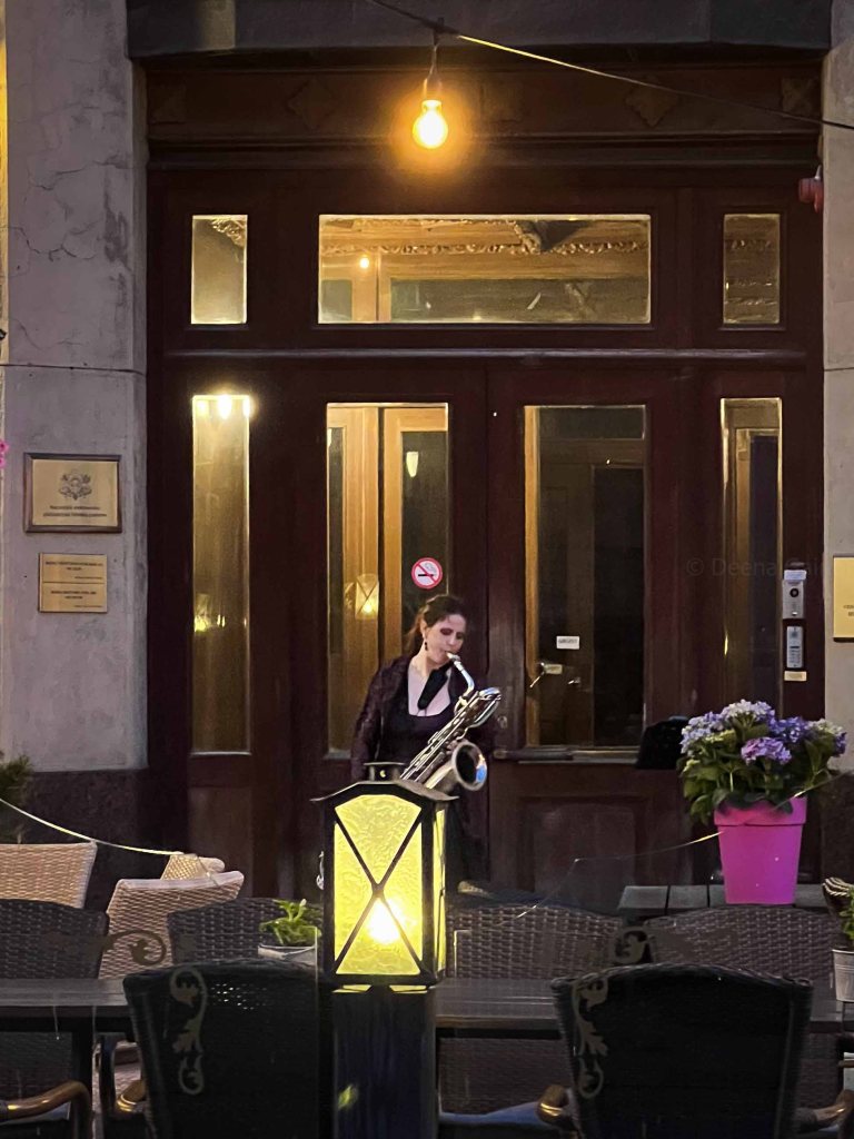 A musician playing a saxophone outside a bar, illuminated by a warm light, with outdoor seating and potted flowers in the foreground.