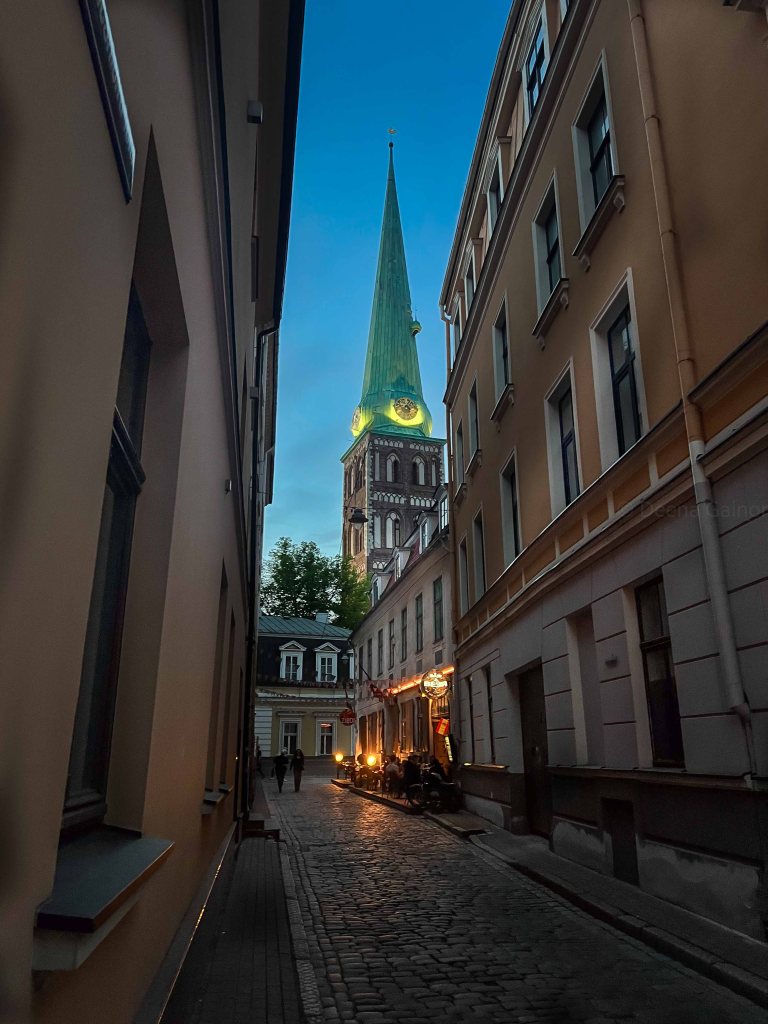 A narrow cobblestone street flanked by tall buildings, leading towards a church tower with a green spire illuminated against a twilight sky.