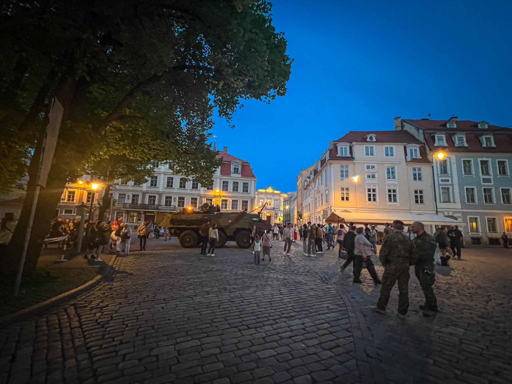 A lively city square at dusk, featuring a military vehicle and a crowd of people, surrounded by historic buildings and streetlights.