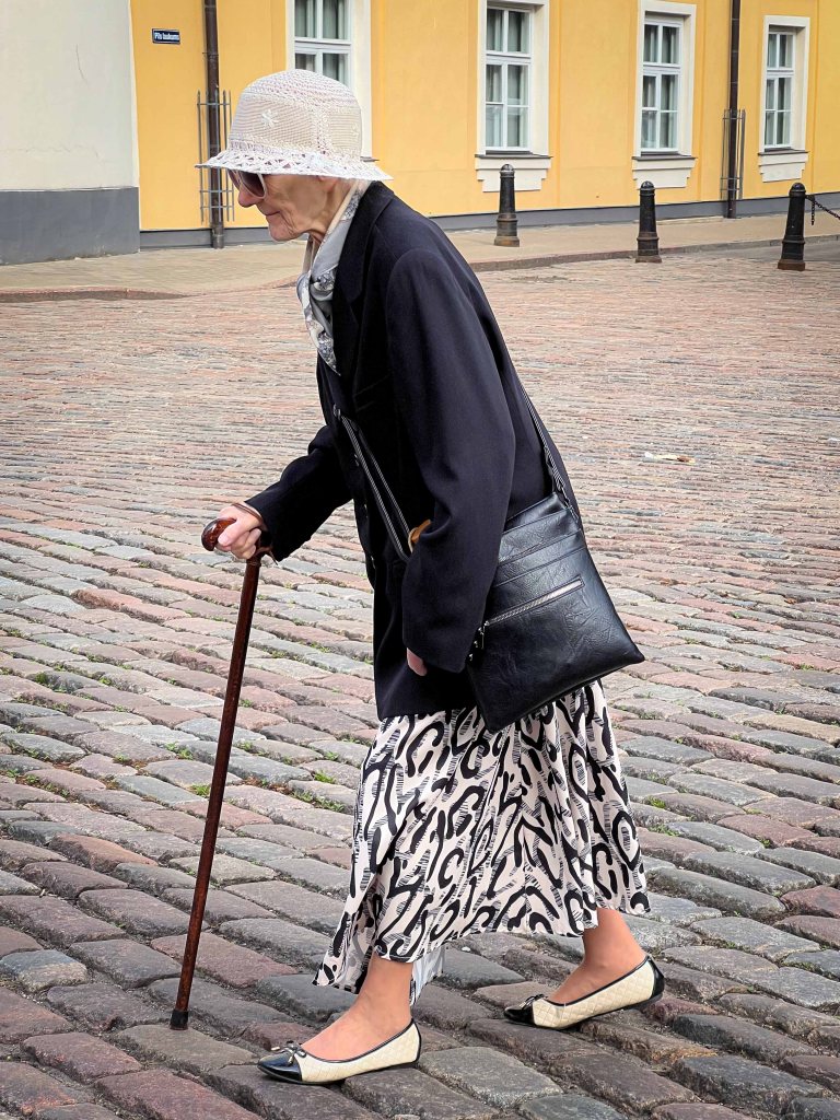 An elderly woman walking with a cane on a cobblestone street, wearing a black coat, a patterned dress, and a wide-brimmed hat, carrying a black handbag.