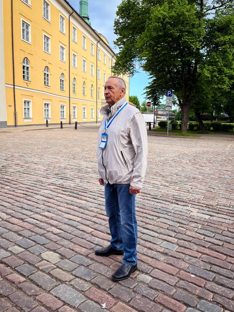 An elderly man stands on a cobblestone street in front of a yellow building, wearing a light jacket and blue jeans.