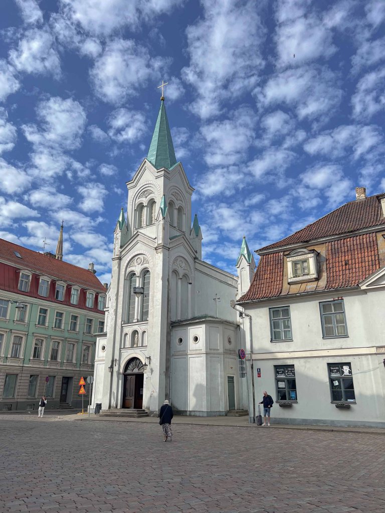 A scenic view of a white church with a green-tipped steeple, surrounded by old buildings and a cobblestone street under a cloudy blue sky.