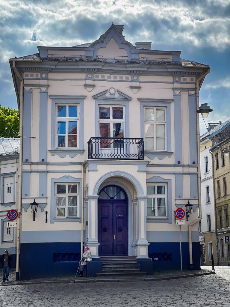 A historic building with a light blue and white façade, featuring a large arched entrance with a purple door, and several windows. A person sits on the steps, while street signs and lamp posts are visible nearby.