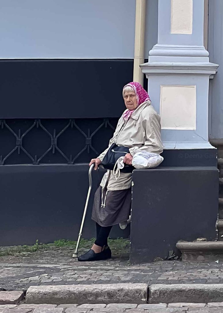 An elderly woman in a pink scarf and gray coat sits against a wall, holding a cane and a bag.