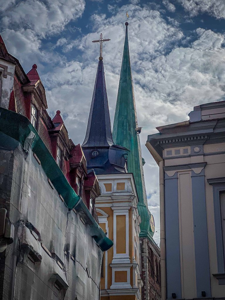 View of two church steeples, one with a green spire and a cross, and the other with a dark, pointed roof, surrounded by historic buildings under a cloudy sky.