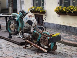 A vintage motorcycle displayed in an outdoor setting, with a rusted frame and flowerpots on the seat and engine.