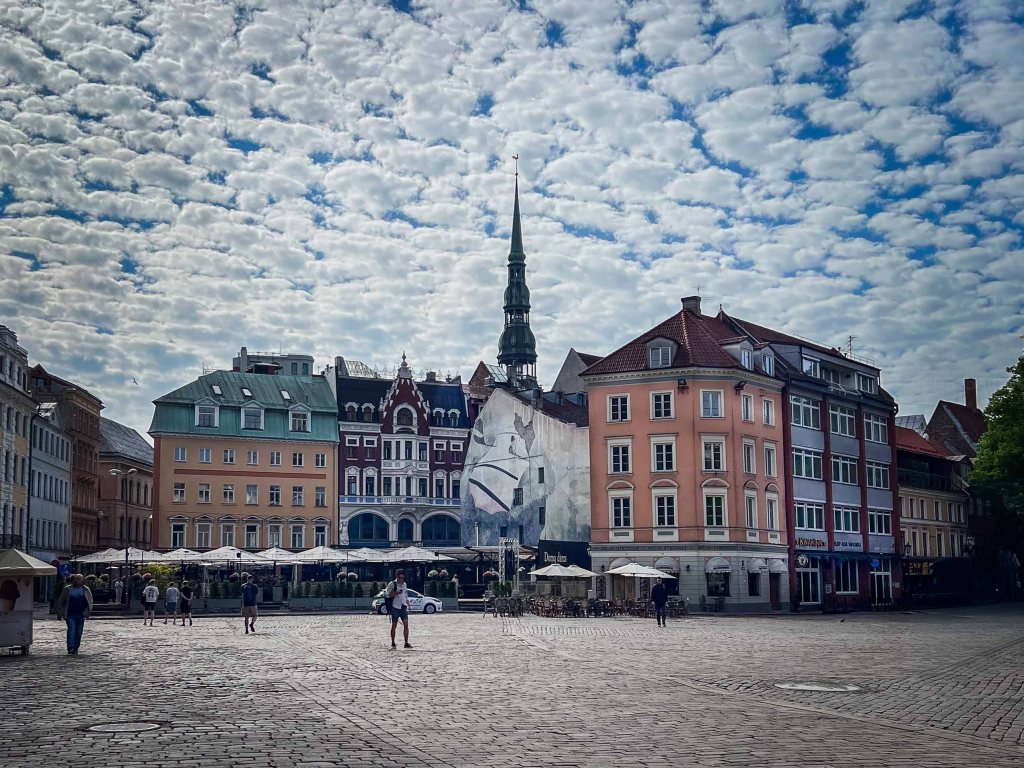 A picturesque square featuring historic buildings with colorful facades and a prominent spire in the background, under a dramatic sky filled with clouds.