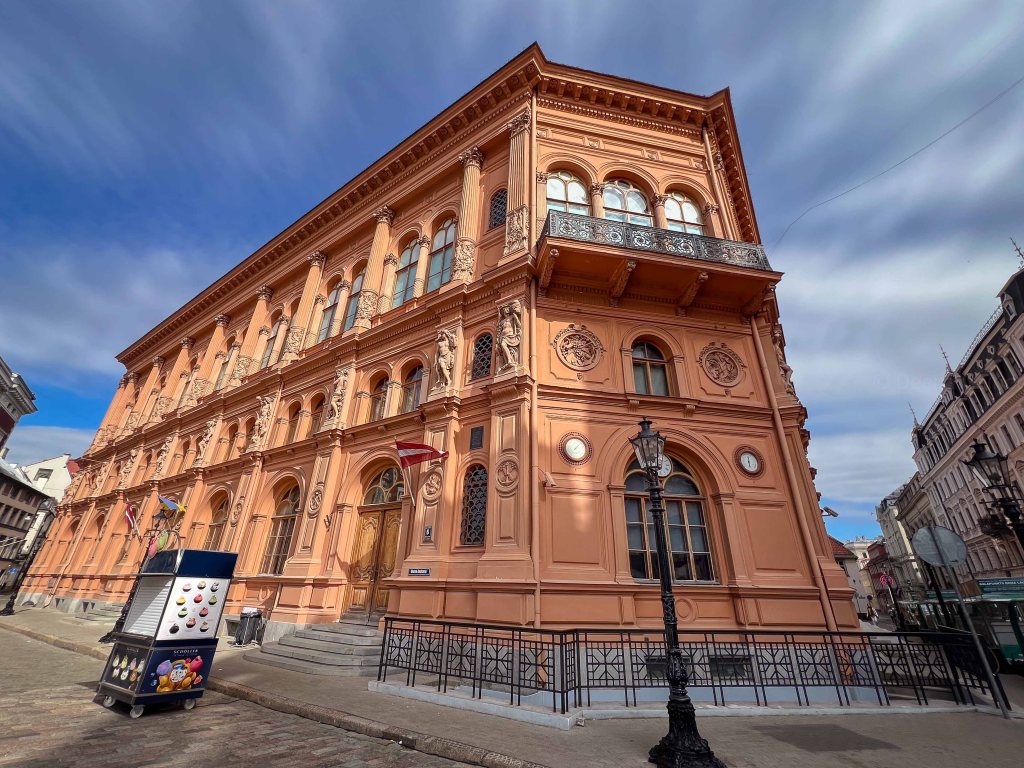 Historic pink building with ornate architecture, featuring large windows and decorative elements, situated on a city street with a parked ice cream cart in the foreground.