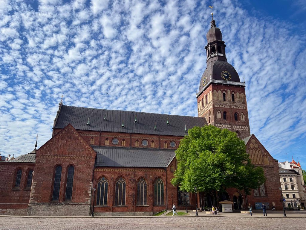 The exterior of a historic brick church with a tall clock tower, surrounded by greenery and a blue sky with clouds.