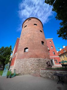Historical brick tower with rounded top, surrounded by a modern building and green trees under a blue sky.