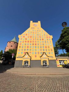 A colorful yellow building with numerous coat of arms displayed on its facade, set against a clear blue sky. In the background, a round tower with a green roof is visible, as well as trees and pavement in the foreground.