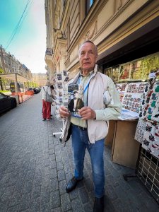 An older man holding a photo while standing on a city street, with art vendors and buildings in the background.