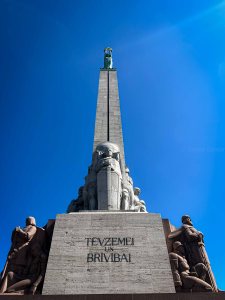 A tall monument featuring a statue at the top, with surrounding figures and inscriptions, set against a clear blue sky.