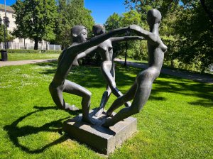 A bronze sculpture depicting three stylized figures in a circular formation, holding hands and engaging in a dance-like pose, set against a green park background.